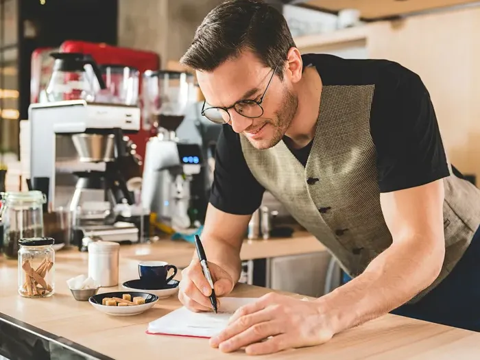 Smiling café owner taking notes at counter with coffee cups and jars, representing local business dedication on townserviceguidenow.com